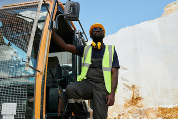 Young African American man in workwear and hardhat standing in excavator or other construction...
