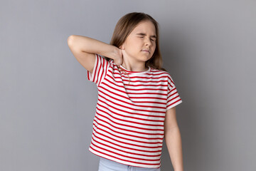 Portrait of unhealthy sick dark haired little girl wearing striped T-shirt standing massaging neck to relieve pain, muscle strain in back. Indoor studio shot isolated on gray background.
