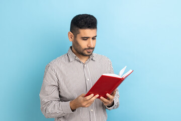 Concentrated bearded businessman holding book and reading, standing absorbed with exciting plot, checking schedule in notepad, wearing striped shirt. Indoor studio shot isolated on blue background.