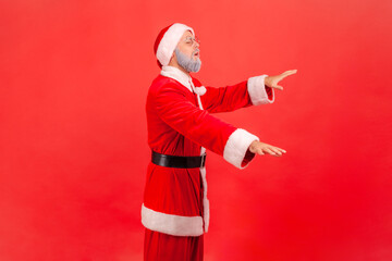 Fototapeta na wymiar Side view of blind disoriented elderly man with gray beard wearing santa claus costume standing with closed eyes and try to touch something. Indoor studio shot isolated on red background.