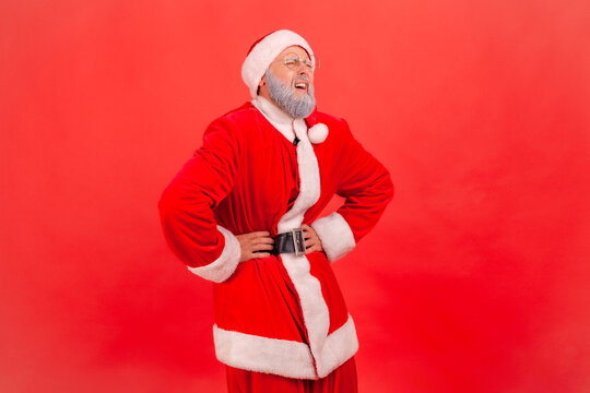 Portrait of elderly man with gray beard wearing santa claus costume standing and keeping hands on belly, suffering from pain, gastritis or constipation. Indoor studio shot isolated on red background.