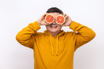 Middle aged man covering eyes with half slices grapefruit and smiling to camera, healthy raw fresh vitamin food concept, wearing urban style hoodie. Indoor studio shot isolated on white background.