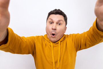 Surprised man taking selfie with shocked facial expression, looking with big eyes and open mouth, wearing urban style hoodie. Indoor studio shot isolated on white background.