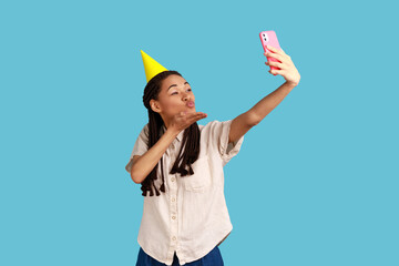 Romantic woman in cone broadcasting livestream, sending air kiss to followers, recording video for blog during celebration birthday, wearing white shirt. Indoor studio shot isolated on blue background