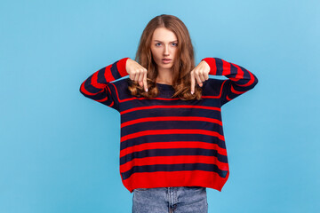 Here and right now. Portrait of boss angry young woman wearing sweater asked for do the task right here and right now, pointing her fingers down. Indoor studio shot isolated on blue background.