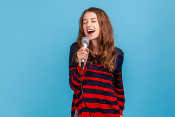 Positive woman wearing striped casual style sweater, holding microphone in hands, singing in karaoke, having fun alone, performing with enthusiasm. Indoor studio shot isolated on blue background.