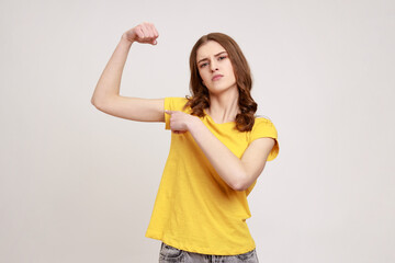 Portrait of proud strong and independent woman of young age in casual style clothing standing and pointing finger at her biceps, showing her power. Indoor studio shot isolated on gray background.
