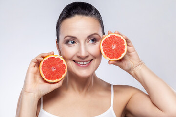 Closeup portrait of middle aged woman smiling and looking away, using balm, mask, lotion for skin care, holding fresh slices grapefruit in her hands. Indoor studio shot isolated on gray background.