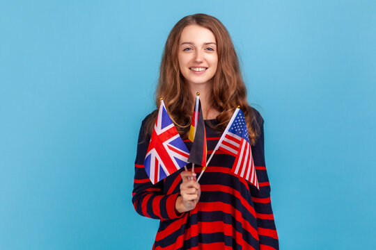 Portrait of young adult woman wearing striped casual style sweater, showing to camera British, American and German flags, expressing happiness. Indoor studio shot isolated on blue background.