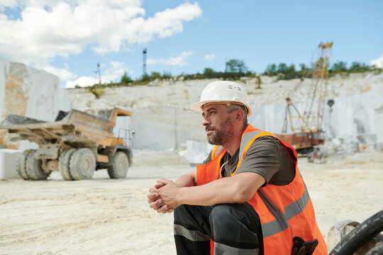 Tired worker of marble quarry sitting in front of camera by workplace against dump truck and working bulldozer with crane
