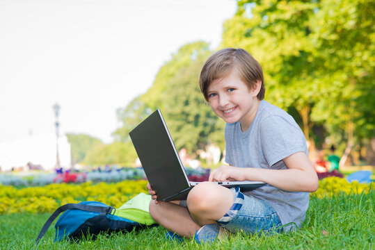 Happy Schoolboy Watching Online Lesson Through Laptop In The Park On The Grass. Handsome Kid Looking For Videos And Other Content On The Internet Outside