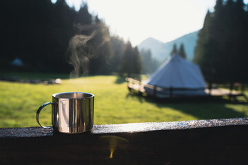 closeup of a steaming metallic cup with warm coffee or tea in the morning with glamping camping ground in the background