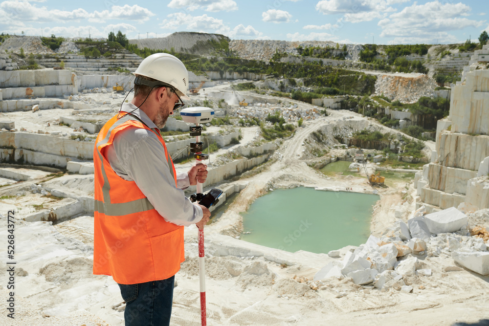 Poster side view of mature engineer or surveyor adjusting theodolite before measuring territory of marble q