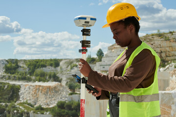 Young serious black woman in workwear and safety helmet adjusting technical equipment for measuring...