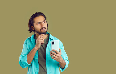 Young pensive casual Indian man with phone doubting looks up and scratches chin when making...