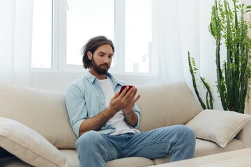 Obraz premium A freelance man in a white T-shirt, blue jeans, and shirt sits on the couch with his phone at home on his day off and plays games
