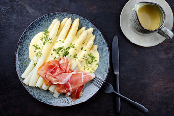 Modern style traditional steamed white asparagus with cured ham and garnished with sauce hollandaise served as top view on a Nordic design plate with copy space