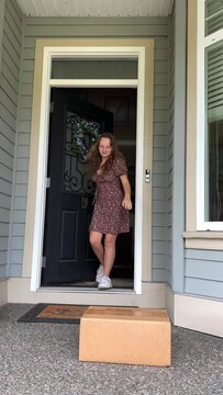 Vertical video where the girls brought the delivery She happily runs out, takes the box, looks around and goes back home She is wearing sneakers and a brown short trendy dress.