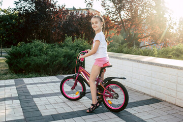 Girl child rides a red bike and is dressed in summer clothes. Happy smiling child with pigtails...