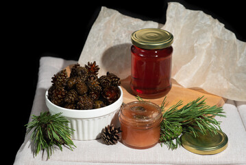 Fototapeta na wymiar On a black background on a light fabric stands delicious caramel jam and syrup from young small pine cones in a glass jar and a bowl with branches and needles