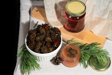 On a black background on a light fabric stands delicious caramel jam and syrup from young small pine cones in a glass jar and a bowl with branches and needles