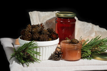 On a black background on a light fabric stands delicious caramel jam and syrup from young small pine cones in a glass jar and a bowl with branches and needles