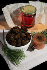 On a black background on a light fabric stands delicious caramel jam and syrup from young small pine cones in a glass jar and a bowl with branches and needles