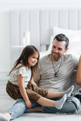 Obraz premium Little cheerful daughter sits on her father and smiles. Happy dad and his little daughter have fun at home. Good relationship between father and daughter. Girl dressed in a skirt and t-shirt
