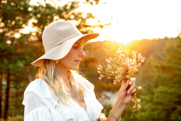 Side view of young beautiful woman standing at sunset in forest, holding, looking at bouquet of...