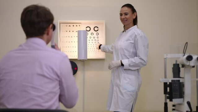 Smiling ophthalmologist pointing at circles on eye test chart talking with man sitting holding occluder. Portrait of expert Caucasian woman checking vision of adult patient in hospital