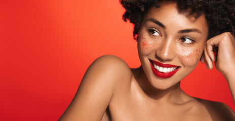 Close up of beautiful african american woman with red lipstick, happy smile, bare glowing shoulders and body, gazing away, standing over studio background