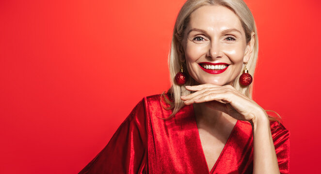 Mature woman with beautiful red lipstick and christmas earring, smiling and looking happy at camera, touching glowing and nourished facial skin after skincare cosmetics