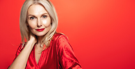 Sensual mature woman of 50 years old, wearing red gown and lipstick, smiling at camera, standing over studio background