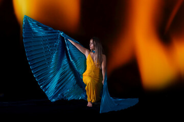 mujer joven vestida de amarillo posando con alas de Isis de color azul , con fondo negro y luces de fuego