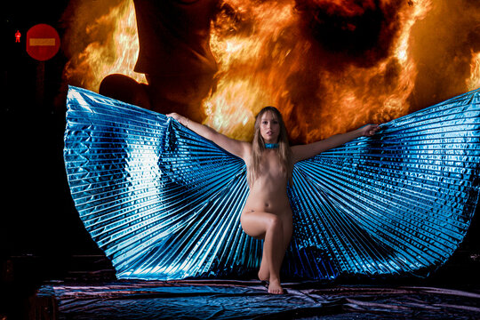 mujer joven posando con alas de Isis de color azul , semidesnuda con fondo negro y luces de fuego