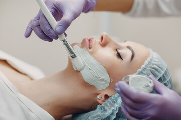 Young woman applying mask of clay on face in beauty spa.