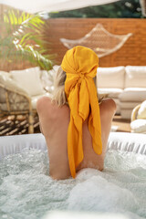 Back view of a woman relaxing in a jacuzzi. Girl wearing fashionable head scarf. Real summer lifestyle, wellness spa concept. Boho terrace at home.