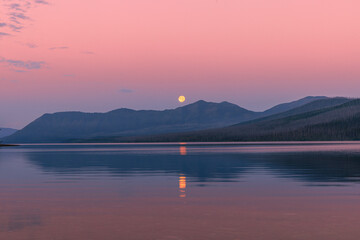Moonset over Lake MacDonald at Glacier National Park, with lake reflections and dawn pinky sky.