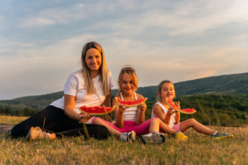Young mother with children outdoor. Cute little caucasian girls and their mother eating watermelon outdoor at hot summer day. Kids siblings spending time together and having fun during summer.