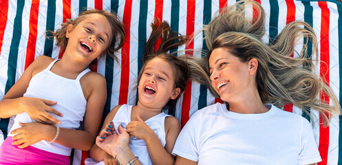 Fototapeta premium Happy mother and daughters smiling and laughing lying on blanket at the lawn in summer park. Family spend their free time together on vacation.