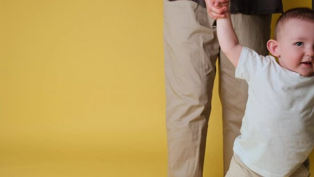 Happy toddler baby learns to walk and takes his first steps with the help mother. Child boy walks with his mom for the first time on a studio yellow background. Kid age one year