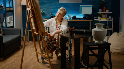 Artist dealing with chronic disability and drawing masterpiece on white canvas, woman with impairment creating artistic masterpiece. Wheelchair user looking at vase to draw sketch.