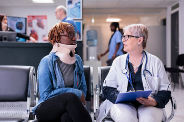 Patient with medical neck collar at consultation with doctor, sitting in waiting room lobby. Woman with cervical foam and senior medic filling in checkup report papers at appointment in hospital.