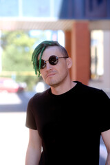 Young stylish man with green dreadlocks in black sunglasses, black t-shirt posing on city street.