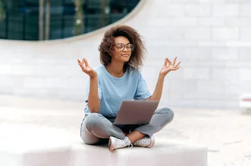 Fotobehang Calm relaxed curly african american young woman with glasses, office worker, sits in a lotus position outdoors near the business center, meditates, relaxes, closed her eyes. Break during online work © Kateryna
