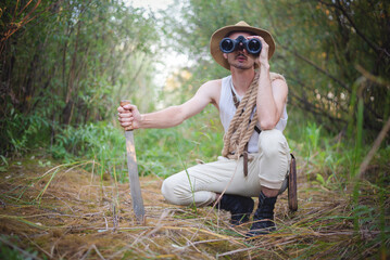 Adventurer man with machete knife is looking through the binoculars in the jungle thicket concept. - obrazy, fototapety, plakaty
