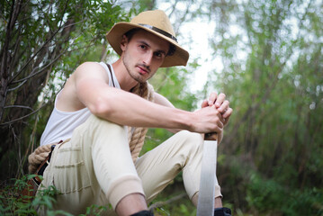 Adventurer man with machete knife sits on the ground in the jungle thicket concept. - obrazy, fototapety, plakaty