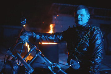 Motorbiker in the black leather jacket with the spikes in the blue light on the burning fire...