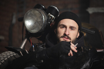 Fototapeta na wymiar Motorbiker in the black leather jacket smokes the cigarette near the the old motorbike concept.
