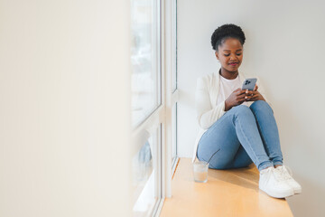 Smiling African girl sitting on a cafe windowsill checking the data on the mobile phone. Afro Hairstyle, enjoying moment, happy people, city life